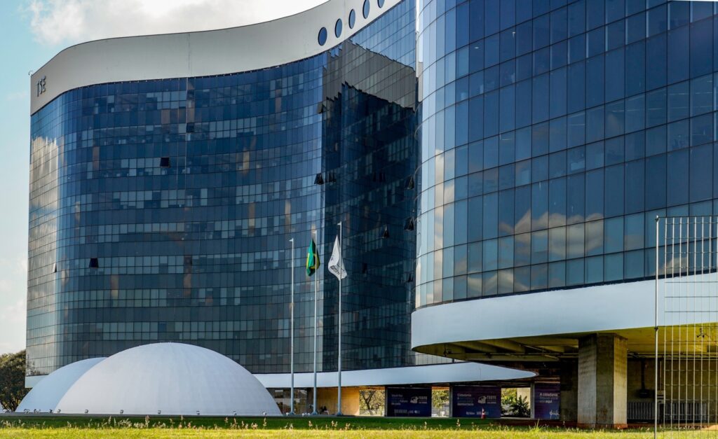 Fachada do edifício do Tribunal Superior Eleitoral com domos brancos ao fundo e a bandeira do Brasil hasteada.