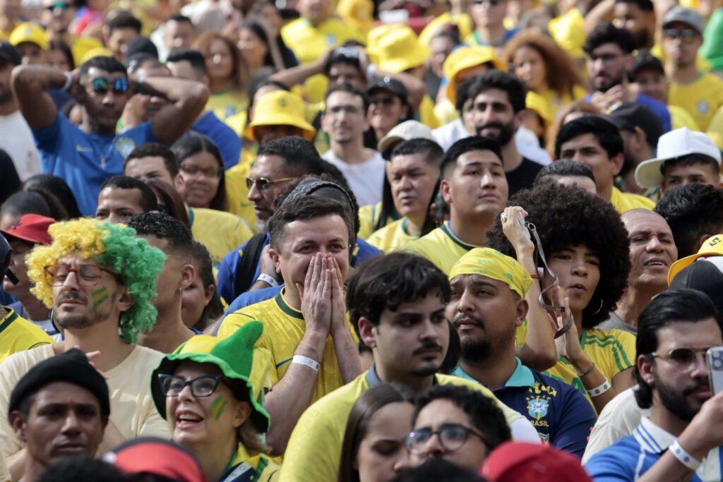 Torcedores com camisas amarelas e adereços do Brasil assistem a um jogo em uma Fan Fest, com expressões de ansiedade e expectativa.