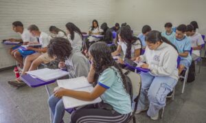 Estudantes do ensino médio sentados em carteiras roxas escrevendo em cadernos em uma sala de aula.