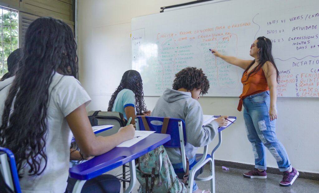 Uma professora aponta para uma lista de verbos em inglês em um quadro branco, enquanto alunos escrevem em seus cadernos em uma sala de aula.