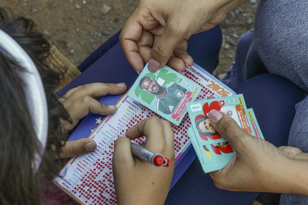 Mãos de pessoas manuseando figurinhas e marcando a lista de países em um álbum com a inscrição "FIGURINHAS 2026".