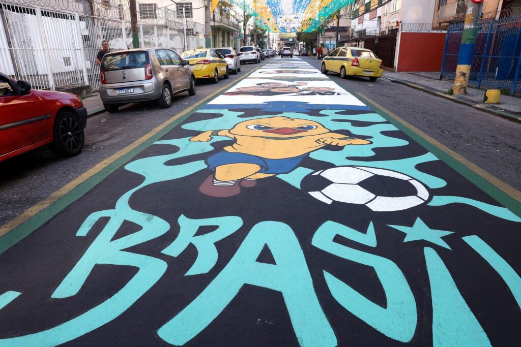 Rua com pinturas coloridas no asfalto, incluindo a palavra "BRASIL" e a imagem de um pato amarelo com uma bola de futebol.