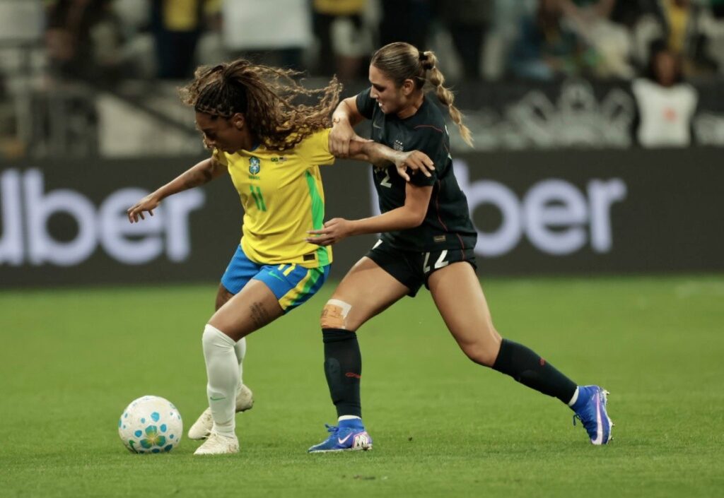 Jogadoras da Seleção Brasileira Feminina e dos Estados Unidos disputam a posse de bola em campo.
