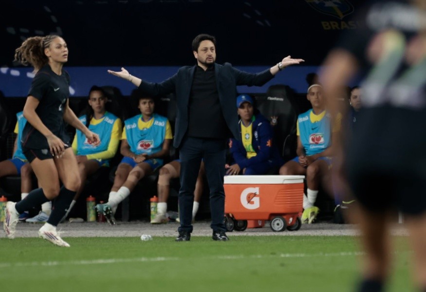 Treinador de terno escuro gesticula com os braços abertos em um campo de futebol, com jogadoras da Seleção Brasileira Feminina ao fundo.