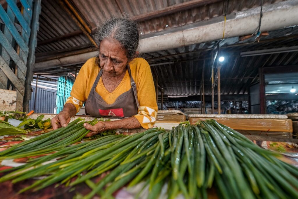 Mulher idosa de blusa amarela e avental organiza maços de cebolinha em uma banca de mercado.