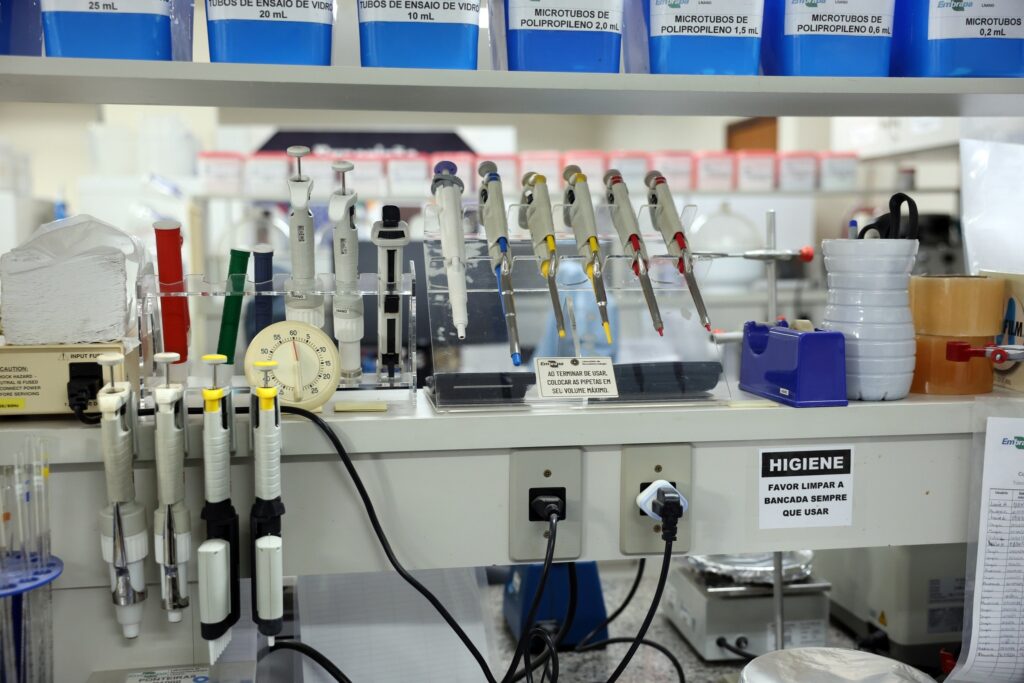 Bancada de laboratório com pipetas, cronômetro e recipientes plásticos azuis. Um aviso na bancada diz: "HIGIENE FAVOR LIMPAR A BANCADA SEMPRE QUE USAR".