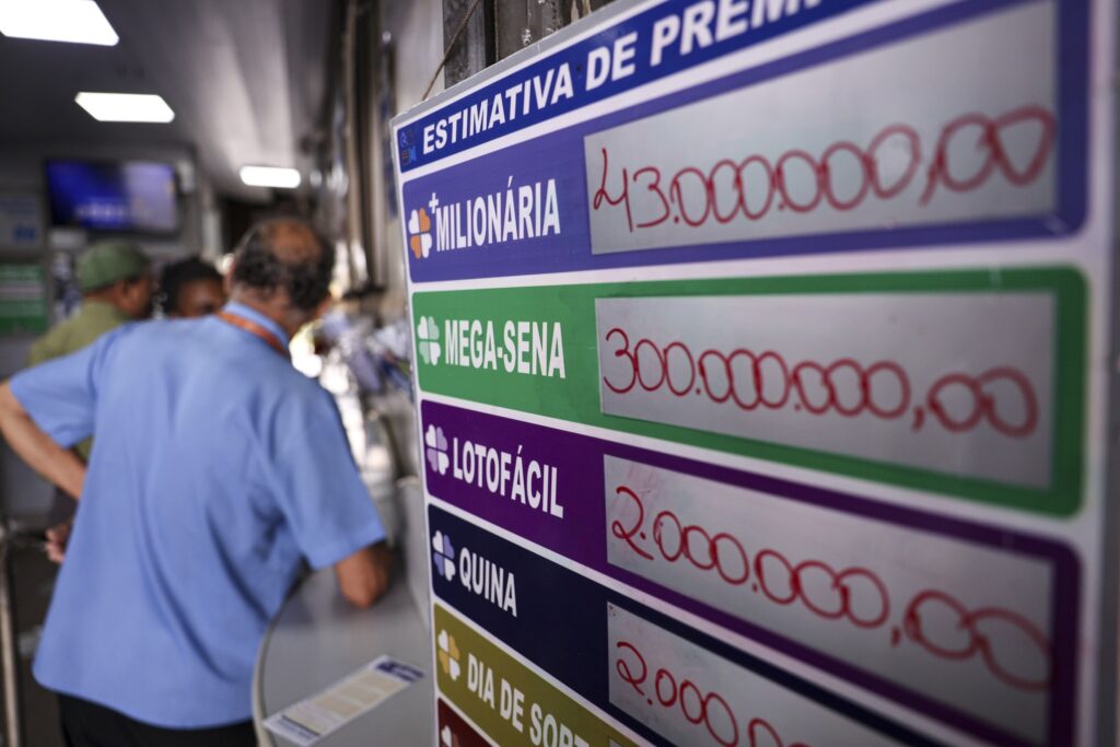Painel de estimativas de prêmios de loterias, destacando a Mega-Sena com o valor "300.000.000,00", enquanto um homem de camisa azul observa a placa ao fundo.