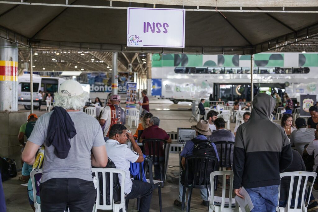 Pessoas aguardam atendimento em um mutirão social sob uma tenda com a placa "INSS" e "Pop Rua Jud", com ônibus ao fundo.