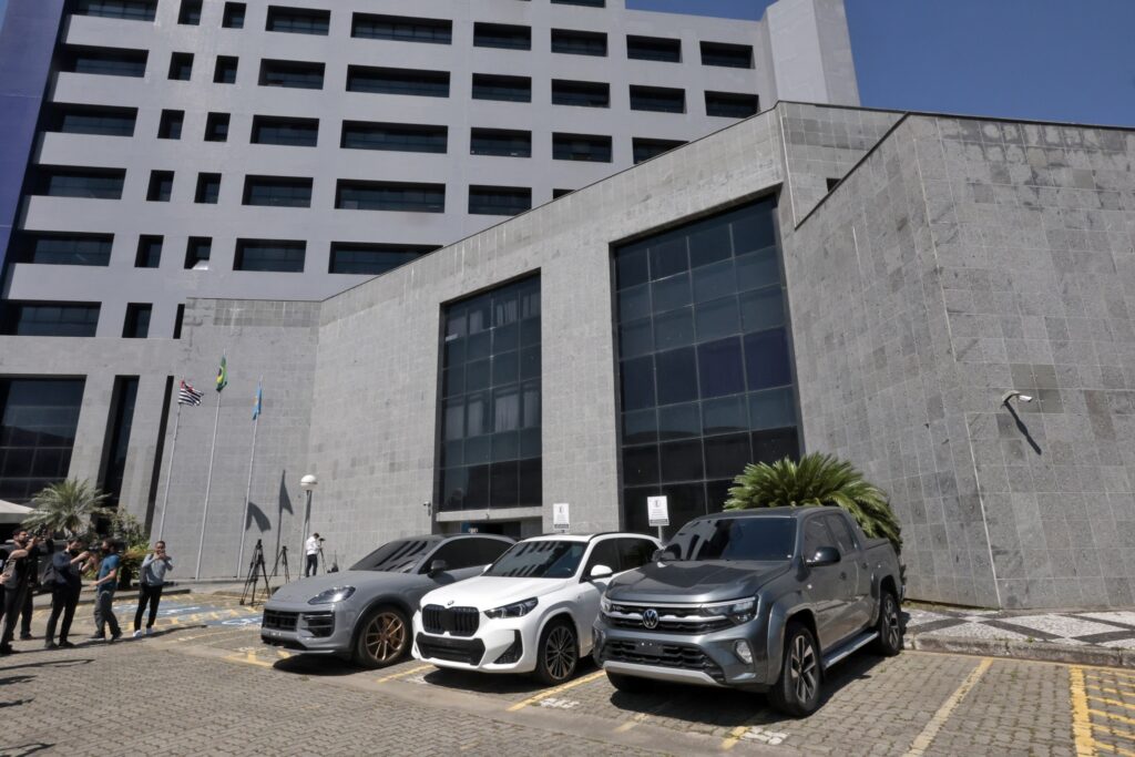 Três carros de luxo, um Porsche, um BMW e uma Volkswagen, estacionados em frente a um prédio institucional durante a "Operação Narcofluxo".