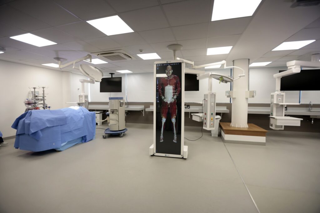 Sala de simulação médica com mesa cirúrgica coberta por pano azul e tela vertical exibindo modelo anatômico do sistema muscular humano.