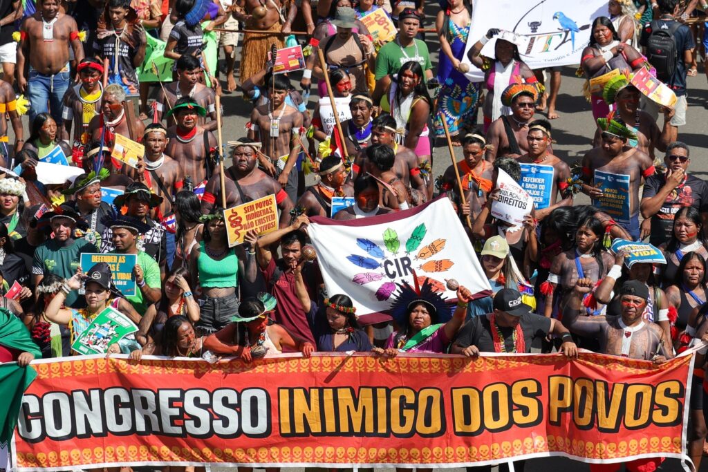 Indígenas em marcha segurando faixa com os dizeres "CONGRESSO INIMIGO DOS POVOS" e diversas placas de reivindicação.