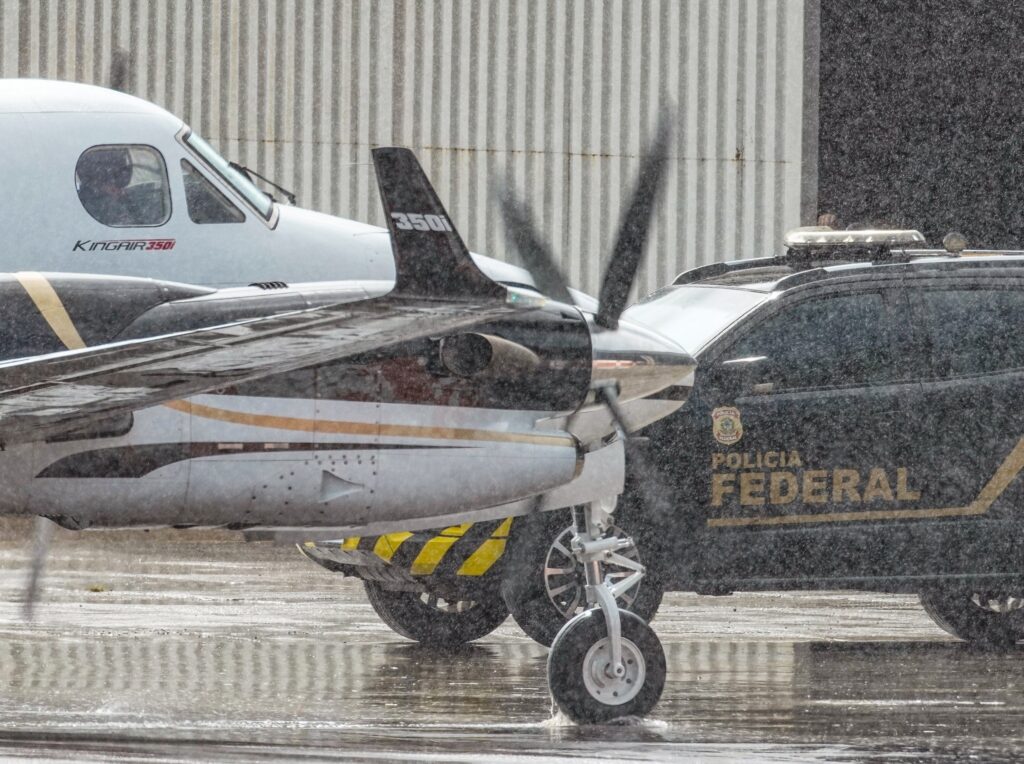 Avião King Air 350i estacionado ao lado de viatura da "POLICIA FEDERAL" sob chuva em pátio molhado.