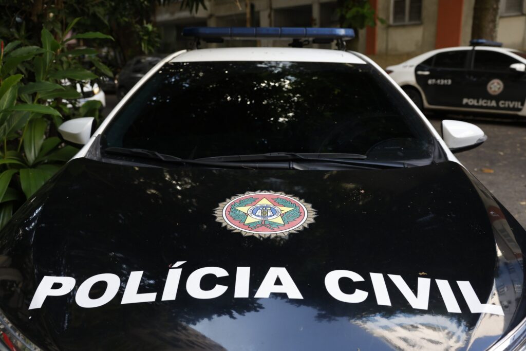 Vista frontal de viatura com a inscrição "POLÍCIA CIVIL" e brasão no capô, com luzes de emergência azuis no teto.