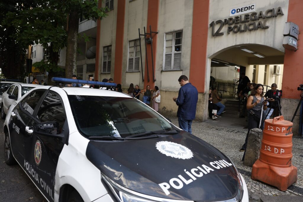 Viatura da Polícia Civil estacionada em frente à "12ª Delegacia Policial", com pessoas reunidas na calçada.
