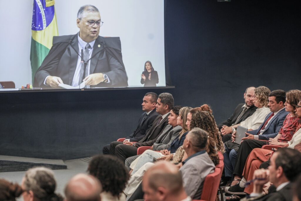 Pessoas em traje formal assistem a uma projeção de um juiz com toga e a imagem de uma mulher durante sessão judicial.