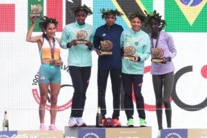 Cinco atletas femininas com coroas de louros posam no pódio com troféus da "100ª Corrida Internacional de São Silvestre".