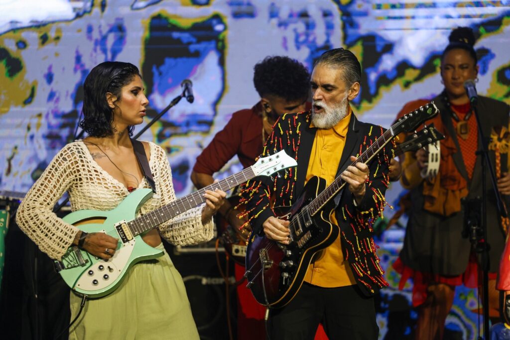 Músicos do grupo Siriará apresentam show com guitarras e performance artística em frente a um painel colorido.