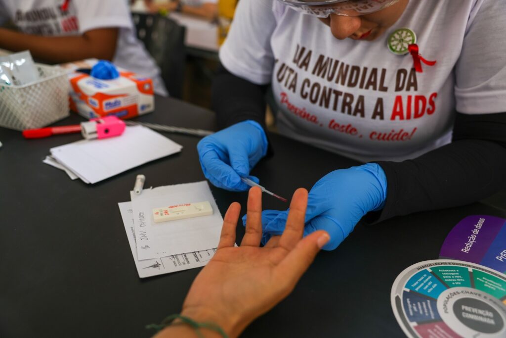 Profissional de saúde com luvas azuis realiza teste rápido de HIV no dedo de uma pessoa. A camiseta do profissional traz a frase "DIA MUNDIAL DA LUTA CONTRA A AIDS".