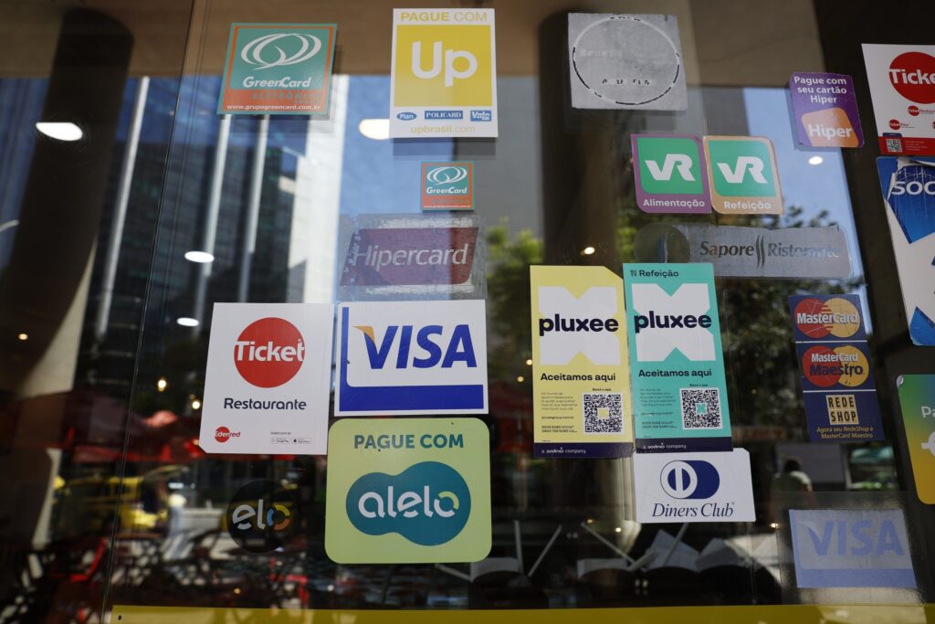 Diversos adesivos de operadoras de cartões de benefícios e pagamentos, como Alelo, Ticket, Visa e VR, colados em uma vitrine de vidro.