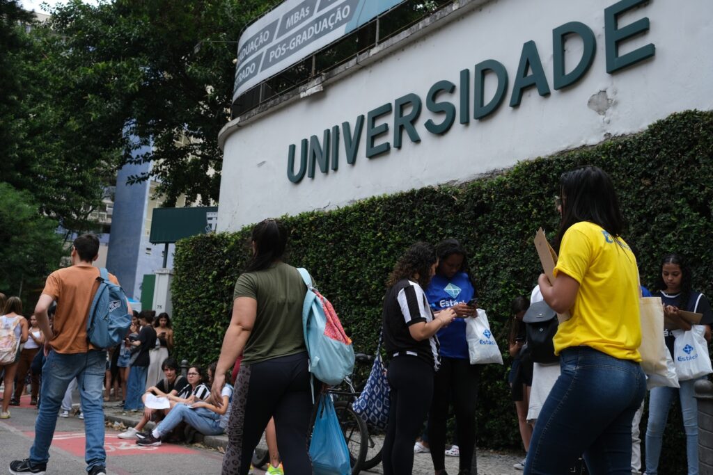 Candidatos aguardam em fila diante de um muro com a palavra 'UNIVERSIDADE' para a realização do Enem.