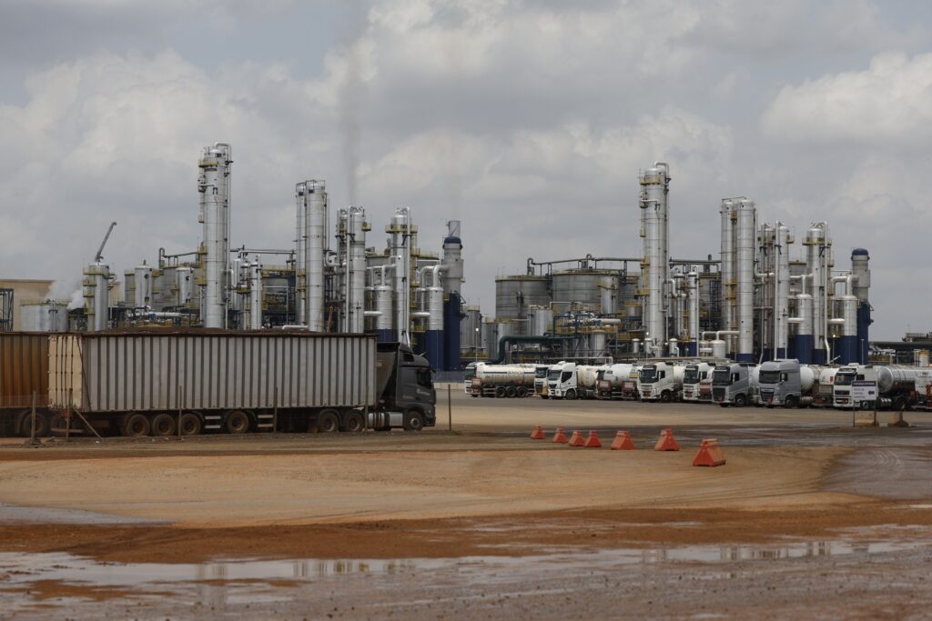 Vista de uma biorefinaria com torres metálicas, tanques e caminhões-tanque estacionados sob um céu nublado.