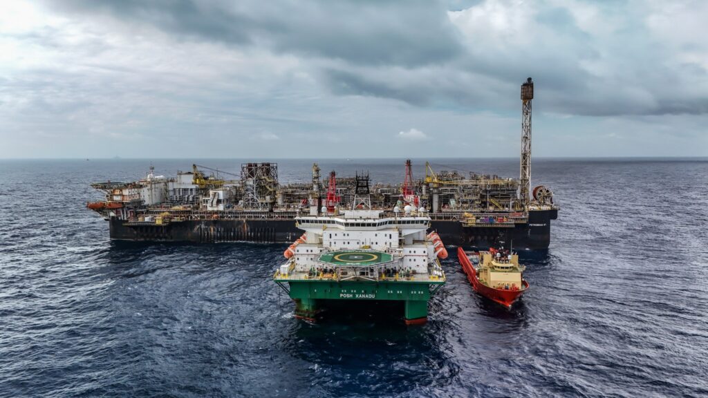 Plataforma de petróleo "PETROBRAS 67" no mar, acompanhada pelo navio de apoio "POSH XANADU" e outra embarcação menor de cor vermelha.