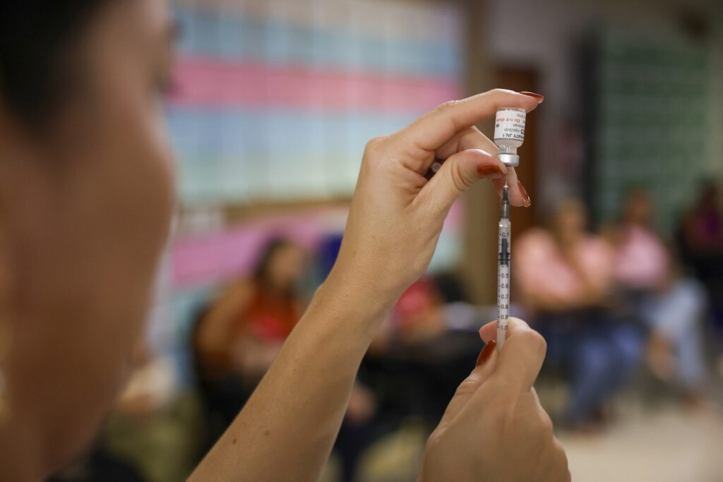 Mãos de profissional de saúde aspirando vacina de um frasco com a inscrição "COMIRNATY JN.1" para uma seringa.