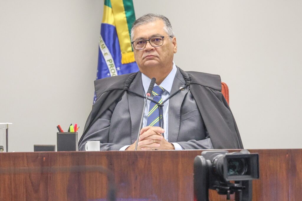 Ministro Flávio Dino, vestindo toga e óculos, sentado atrás de bancada de madeira em plenário, com a bandeira do Brasil ao fundo.