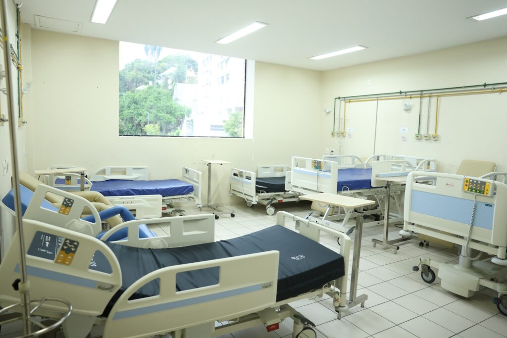 Interior de uma enfermaria hospitalar com várias camas articuladas com colchões azuis e mesas de apoio.