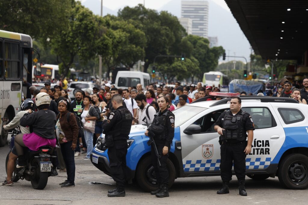 Três policiais militares ao lado de uma viatura branca e azul em frente a uma multidão de pessoas em uma rua urbana.