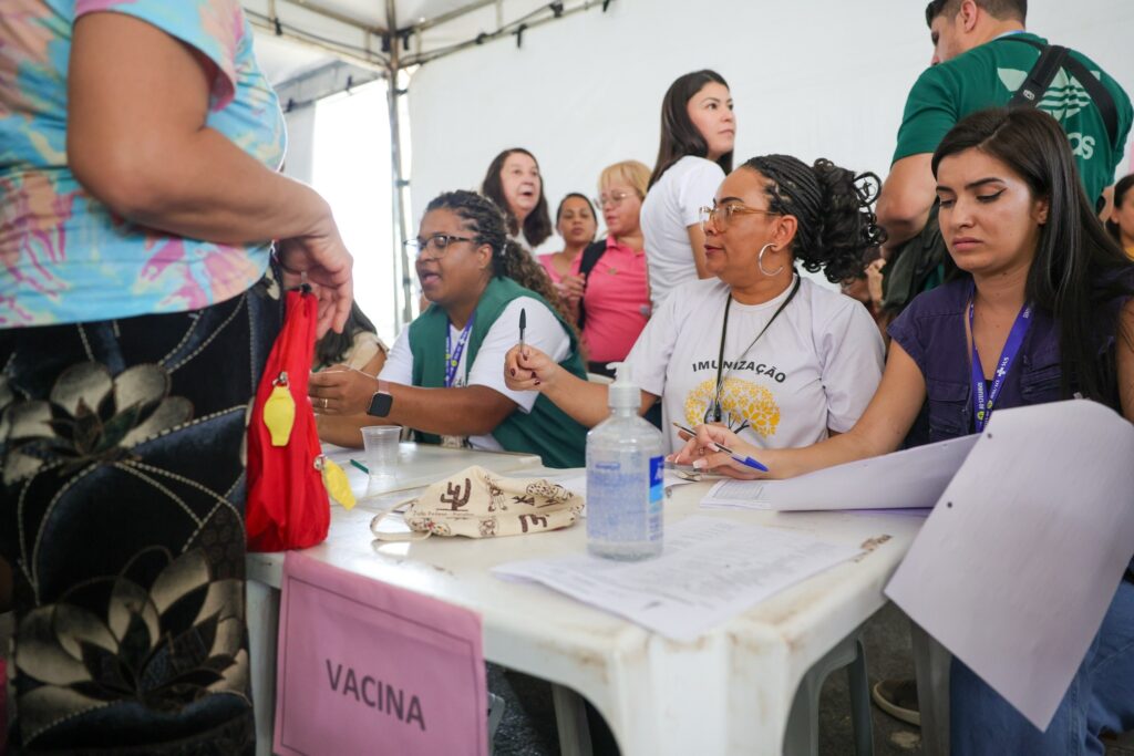 Profissionais de saúde atendem em uma mesa com a placa "VACINA" e camiseta com a escrita "IMUNIZAÇÃO" durante campanha de vacinação.