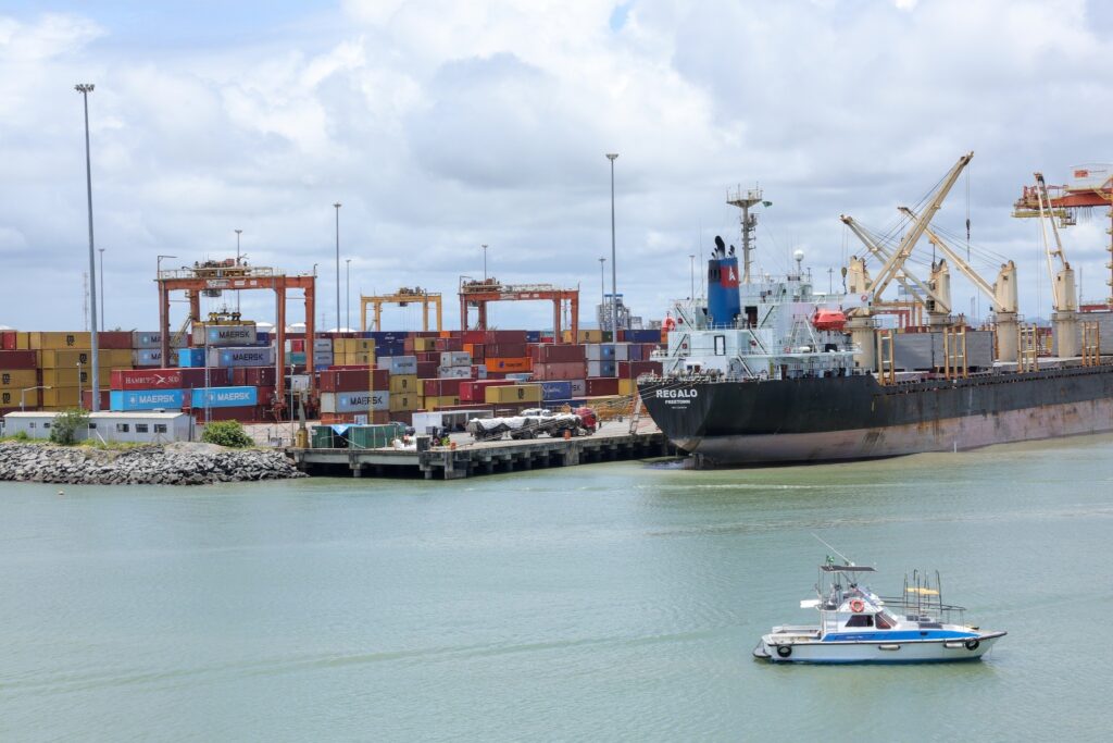 Vista do Porto de Suape com um grande navio cargueiro atracado, containers empilhados ao fundo e um pequeno barco branco navegando na água.