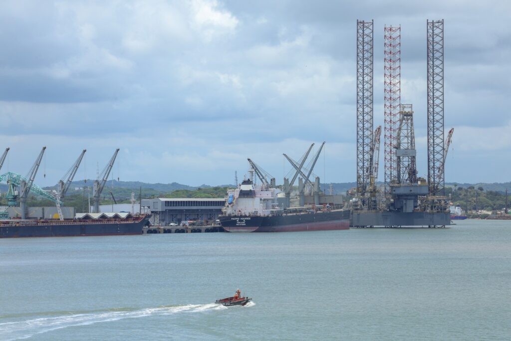 Vista do Porto de Suape com navios de carga, guindastes e uma plataforma de petróleo, com uma pequena embarcação navegando em primeiro plano.