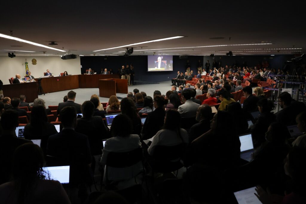 Público sentado em cadeiras vermelhas assiste a uma sessão no Supremo Tribunal Federal, com a imagem de um homem em um telão ao fundo.