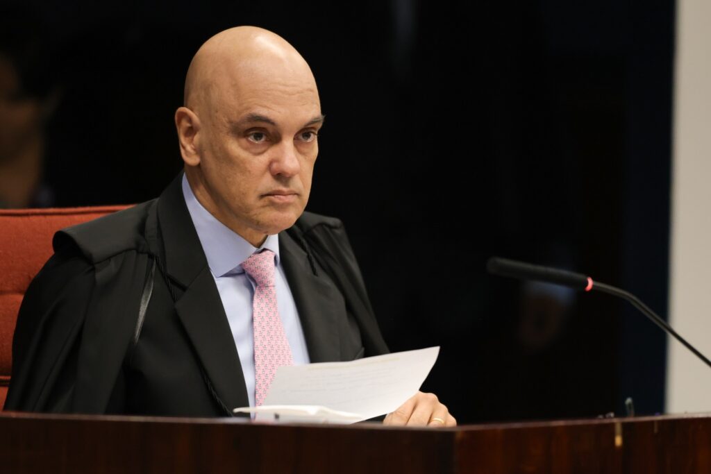 Ministro Alexandre de Moraes, trajando toga e gravata rosa, segura papéis durante sessão do Supremo Tribunal Federal.