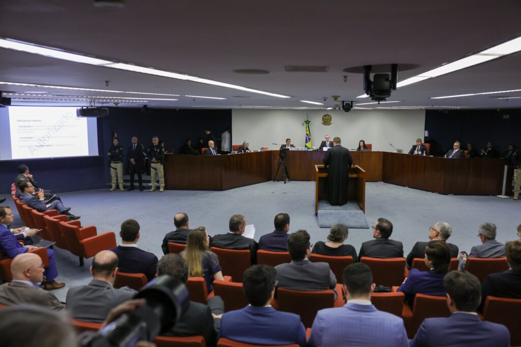 Sessão de julgamento no STF com público em poltronas laranjas e orador de toga. Uma tela exibe o texto "DA IMPERIOSA ABSOLVIÇÃO DO GENERAL DELENIO".