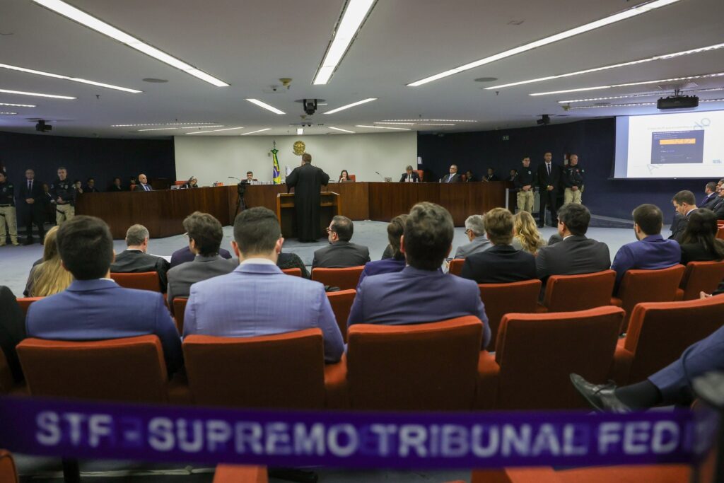 Pessoas assistem a um julgamento no STF, com uma pessoa de toga diante da bancada de juízes e a faixa "STF - SUPREMO TRIBUNAL FED" em primeiro plano.