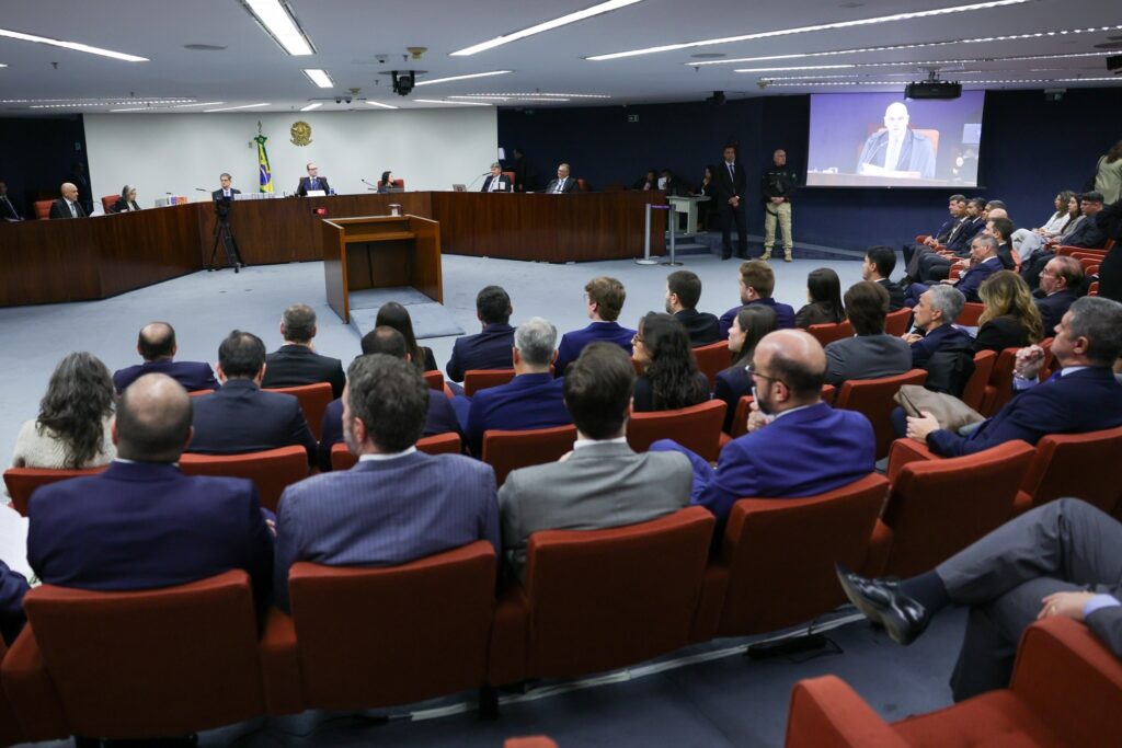 Pessoas sentadas em poltronas vermelhas assistem a um julgamento em um tribunal, com magistrados em uma bancada ao fundo e a imagem de um juiz em um telão.