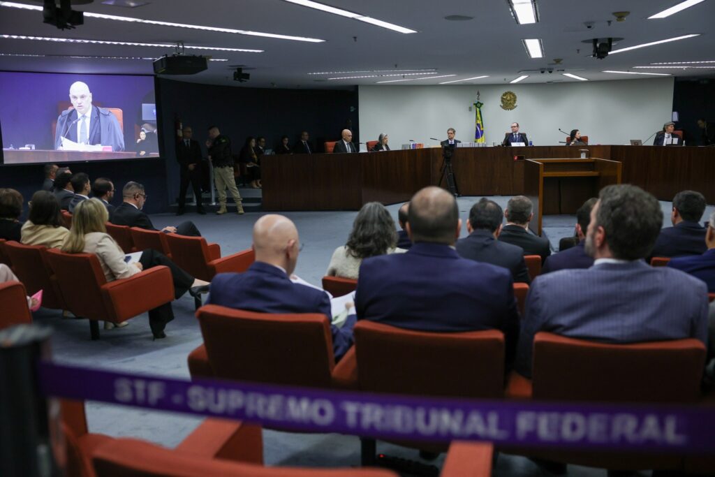 Pessoas sentadas em poltronas vermelhas assistem a uma sessão no Supremo Tribunal Federal, com a imagem de um homem falando em um telão ao fundo e a inscrição "STF - SUPREMO TRIBUNAL FEDERAL" em uma fita no primeiro plano.