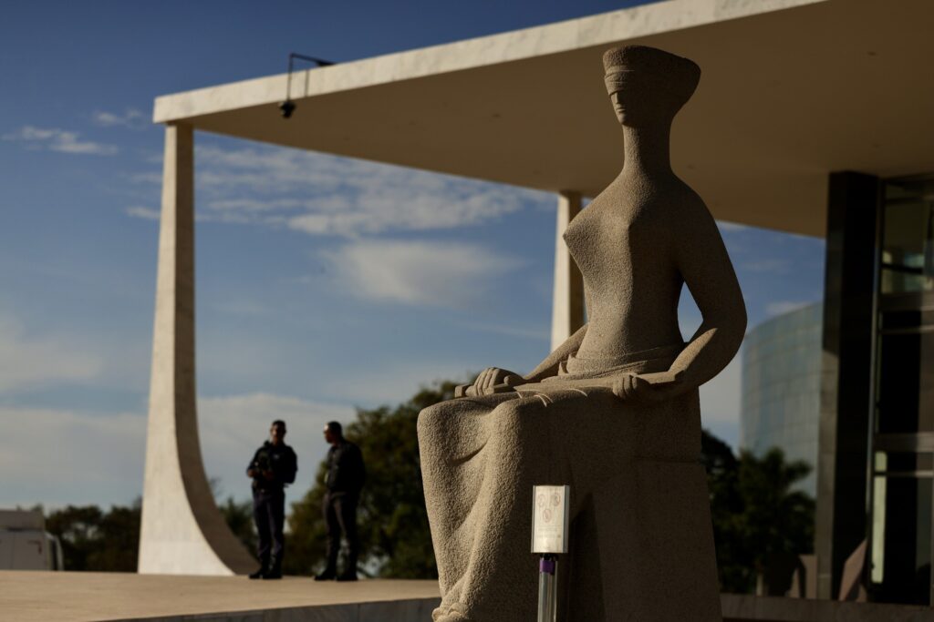 Estátua da Justiça, com venda nos olhos, em frente ao Supremo Tribunal Federal, com agentes de segurança ao fundo.