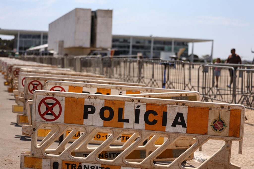 Barreiras de segurança com a palavra "POLÍCIA" em primeiro plano, com grades de contenção e prédios governamentais ao fundo.