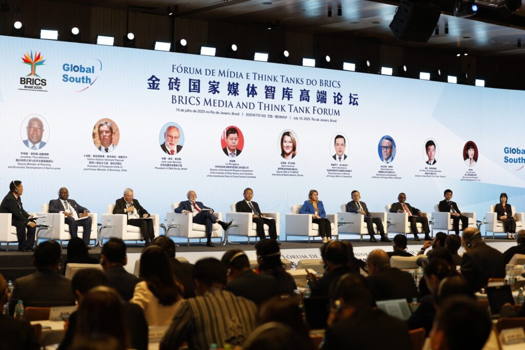 Painel de discussão no "Fórum de Mídia e Think Tanks do Brics", com onze pessoas sentadas em poltronas brancas diante de um telão com logotipos e retratos de palestrantes.