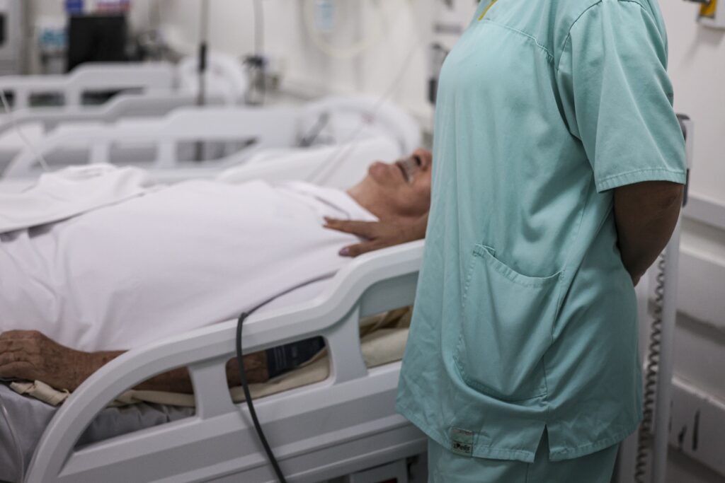 Profissional de saúde ao lado de paciente em leito de hospital.