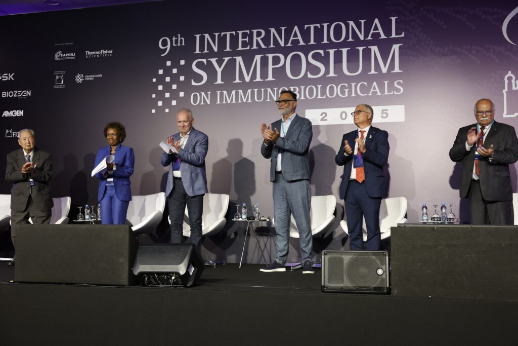 Seis pessoas em trajes formais aplaudem no palco diante de um painel com os dizeres "9th INTERNATIONAL SYMPOSIUM ON IMMUNOBIOLOGICALS 2025".