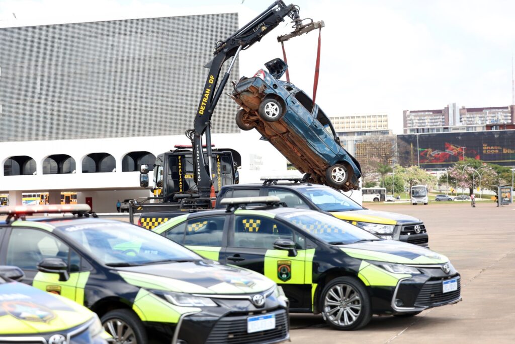 Um guincho do Detran-DF suspende um carro azul destruído sobre viaturas de fiscalização em uma praça, como parte da campanha "Maio Amarelo".
