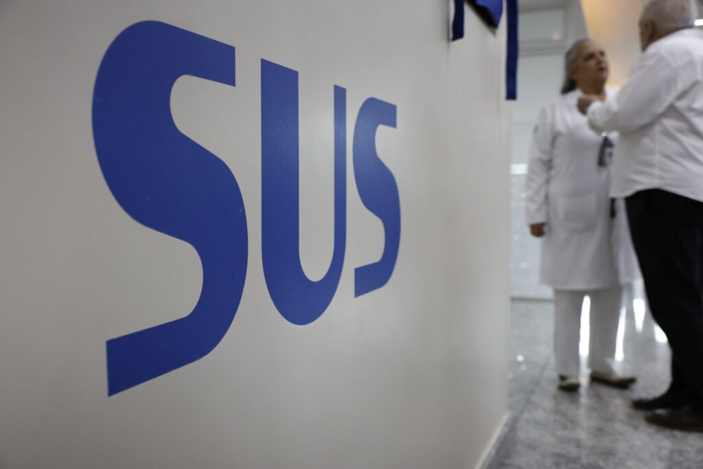 Logotipo "SUS" em destaque em uma parede branca, com profissionais de saúde desfocados ao fundo.