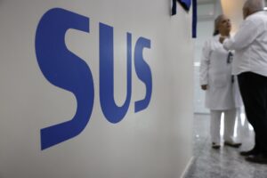 Logotipo "SUS" em destaque em uma parede branca, com profissionais de saúde desfocados ao fundo.