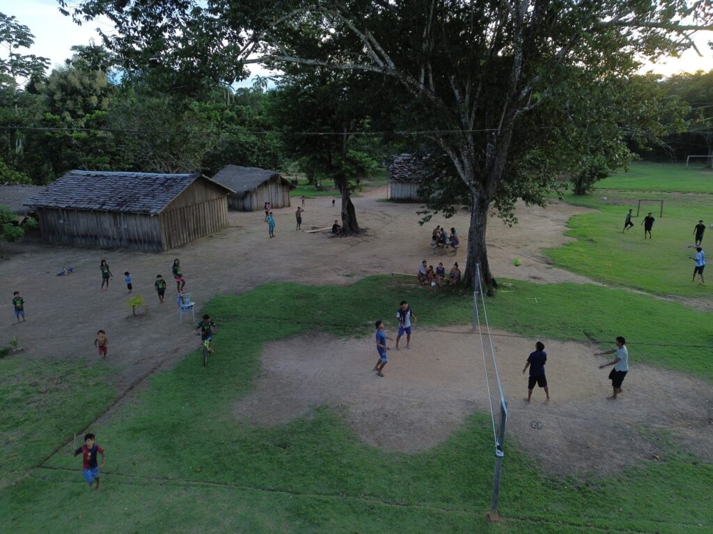 Crianças e adultos praticam vôlei e futebol em uma área aberta de aldeia indígena com casas de madeira e árvores.