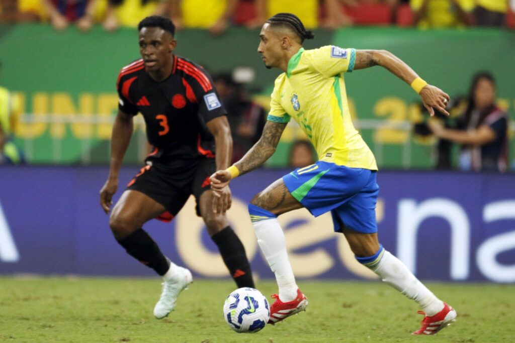 Jogador da seleção brasileira conduz a bola enquanto é marcado por jogador da seleção colombiana em partida de futebol.