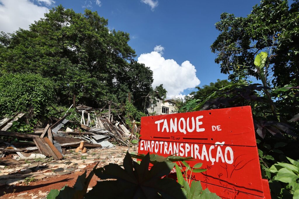 Placa vermelha com os dizeres "TANQUE DE EVAPOTRANSPIRAÇÃO" em frente a um amontoado de madeiras e entulhos em área arborizada.